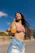 A smiling woman holding a chilled bottle of Frutyka mango juice against a sunny outdoor backdrop.