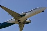A commercial airplane with the logo 'WestJet' on the fuselage is captured in mid-flight against a clear blue sky. The aircraft is white with teal accents on the engines and tail.