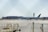 A commercial airplane belonging to Air Canada is on a runway or taxiway at an airport. The background includes hangars or terminal buildings and an air traffic control tower. The foreground shows some airport equipment, likely for ground services.