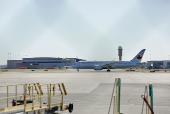 A commercial airplane belonging to Air Canada is on a runway or taxiway at an airport. The background includes hangars or terminal buildings and an air traffic control tower. The foreground shows some airport equipment, likely for ground services.