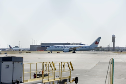 An Air Canada airplane is stationed on an airport tarmac, with airport buildings and a control tower visible in the background. The scene includes airport equipment and a clear sky.