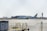 An Air Canada airplane is stationed on an airport tarmac, with airport buildings and a control tower visible in the background. The scene includes airport equipment and a clear sky.