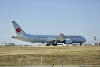 A large commercial airplane with a red maple leaf on its tail is on a runway at an airport. The plane is marked with 'Air Canada' along its side and appears to be either preparing for takeoff or recently landed. The runway is bordered by a grassy area, and an airport terminal with various structures, including a control tower, is visible in the background. The sky is clear, creating a calm and serene ambiance.