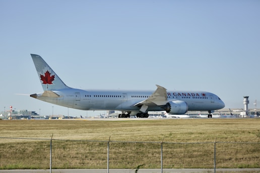 A large commercial airplane with a red maple leaf on its tail is on a runway at an airport. The plane is marked with 'Air Canada' along its side and appears to be either preparing for takeoff or recently landed. The runway is bordered by a grassy area, and an airport terminal with various structures, including a control tower, is visible in the background. The sky is clear, creating a calm and serene ambiance.