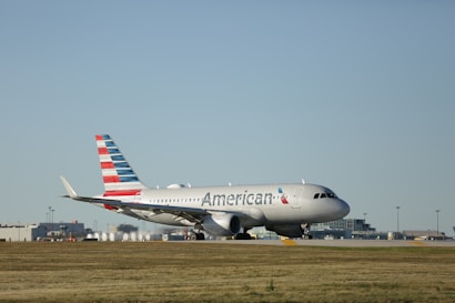 A commercial airplane with the logo and design of an airline is parked on an airport runway. The aircraft is white with a distinctive red, white, and blue tail fin. It is situated on a flat stretch of grass with airport buildings visible in the background under a clear blue sky.