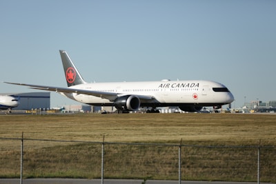 An Air Canada aircraft positioned on a runway at an airport, with buildings and infrastructure visible in the background under a clear sky.