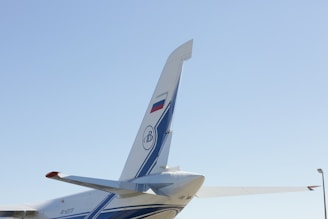 The image features the tail section of an aircraft with a visible fin and rudder, adorned with blue and white stripes and a logo. The tail displays a national flag and is set against a clear blue sky. Part of the horizontal stabilizer is visible alongside the tail.