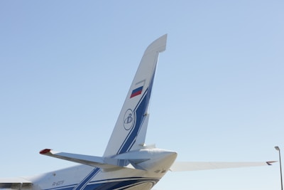 The image features the tail section of an aircraft with a visible fin and rudder, adorned with blue and white stripes and a logo. The tail displays a national flag and is set against a clear blue sky. Part of the horizontal stabilizer is visible alongside the tail.