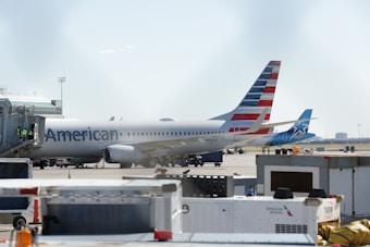 A commercial airplane is parked at an airport gate, with a jet bridge attached. The American Airlines logo is visible on the aircraft's body, while several airport workers are seen wearing safety vests near the jet bridge. The tail fin displays the airline's distinctive red, white, and blue stripes. Nearby, there is another plane from a different airline. Various ground equipment and vehicles are scattered around the tarmac.