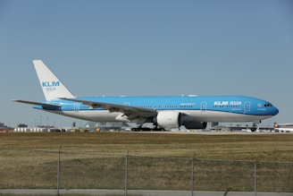 A large commercial airplane with a blue and white color scheme is taxiing on a runway. The aircraft is branded with KLM Asia livery on its fuselage and tail. The background shows airport infrastructure including terminal buildings and other airplanes.