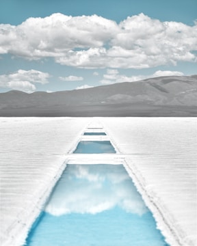 A vast, white salt flat stretches towards the horizon. A narrow, artificial canal filled with clear, reflective water cuts through the landscape, mirroring the partly cloudy sky above. Distant mountains provide a dark contrast to the bright salt and vivid blue sky.