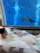 A person is reclining in a bubble-filled bathtub with a large aquarium window in front. Beyond the glass, marine life including fish and a large stingray is visible swimming in the blue water.