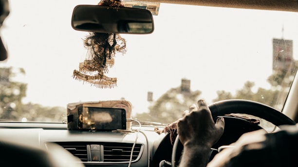 The interior of a car is shown from the perspective of the backseat. There is a driver gripping the steering wheel, with a phone mounted on the dashboard. Ornate decorations are hanging from the rearview mirror. The surroundings outside the car appear blurred, indicating motion.