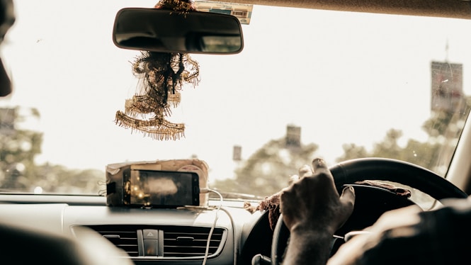 The interior of a car is shown from the perspective of the backseat. There is a driver gripping the steering wheel, with a phone mounted on the dashboard. Ornate decorations are hanging from the rearview mirror. The surroundings outside the car appear blurred, indicating motion.
