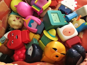 A collection of colorful cartoon figurines arranged randomly. The figures include a variety of characters with different facial expressions and accessories. The figurines are made of plastic and feature playful designs and bright colors.