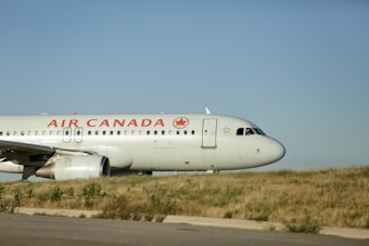 An Air Canada airplane is taxiing on the runway, with the company logo visible on the fuselage. The background shows a clear blue sky and grassy terrain.