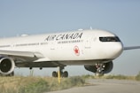 A large, white aircraft with the Air Canada logo and branding is parked on a runway with green grass visible on the sides. The plane is positioned slightly to one side, showcasing its engines and landing gear. The sky is clear and blue, providing a serene backdrop to the scene.