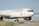 A large, white aircraft with the Air Canada logo and branding is parked on a runway with green grass visible on the sides. The plane is positioned slightly to one side, showcasing its engines and landing gear. The sky is clear and blue, providing a serene backdrop to the scene.