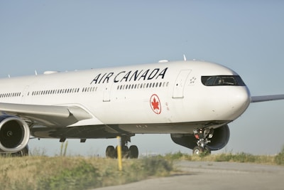 A large, white aircraft with the Air Canada logo and branding is parked on a runway with green grass visible on the sides. The plane is positioned slightly to one side, showcasing its engines and landing gear. The sky is clear and blue, providing a serene backdrop to the scene.