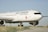 A large, white aircraft with the Air Canada logo and branding is parked on a runway with green grass visible on the sides. The plane is positioned slightly to one side, showcasing its engines and landing gear. The sky is clear and blue, providing a serene backdrop to the scene.