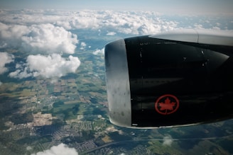 An aerial view of a landscape with scattered clouds and visible farmlands and urban areas below. The image is dominated by the airplane engine, which features an emblem with a red maple leaf.