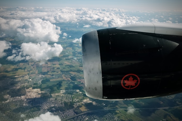 An aerial view of a landscape with scattered clouds and visible farmlands and urban areas below. The image is dominated by the airplane engine, which features an emblem with a red maple leaf.