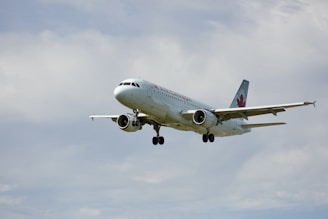 A commercial airplane is flying in the sky with landing gear extended. The aircraft is a white passenger jet with branding and a maple leaf logo on the tail. It is set against a backdrop of a cloudy sky.