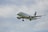 A commercial airplane is flying in the sky with landing gear extended. The aircraft is a white passenger jet with branding and a maple leaf logo on the tail. It is set against a backdrop of a cloudy sky.