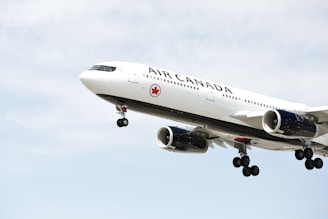 A large Air Canada airplane is in mid-flight with its landing gear extended against a backdrop of a cloudy sky. The aircraft has a sleek white body with the airline's logo visible on the side.