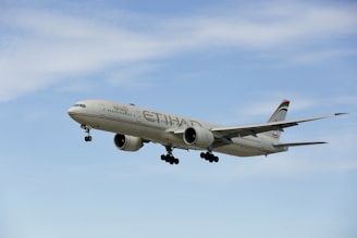 A large commercial airplane with the Etihad Airways branding is flying against a clear blue sky. The airplane is a twin-engine jet with landing gear extended, suggesting it's either approaching for landing or taking off.