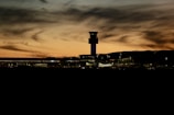 A silhouette of an airport with a control tower at sunset. The sky is filled with swirling clouds in shades of orange and yellow, creating a dramatic backdrop. The dark outlines of the terminal and parked airplanes are visible, along with soft, ambient lighting from the buildings.