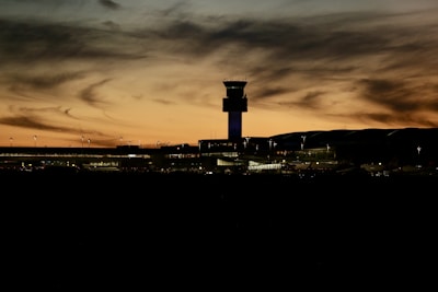 A silhouette of an airport with a control tower at sunset. The sky is filled with swirling clouds in shades of orange and yellow, creating a dramatic backdrop. The dark outlines of the terminal and parked airplanes are visible, along with soft, ambient lighting from the buildings.