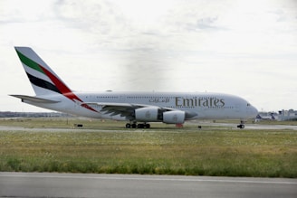 A large Emirates passenger airplane is taxiing on a runway at an airport. The aircraft is painted white with distinctive markings on the tail, featuring red, green, and black stripes. The surrounding area includes grass fields and some distant airport buildings under a cloudy sky.