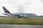 A large Emirates passenger airplane is taxiing on a runway at an airport. The aircraft is painted white with distinctive markings on the tail, featuring red, green, and black stripes. The surrounding area includes grass fields and some distant airport buildings under a cloudy sky.