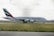 A large Emirates passenger airplane is taxiing on a runway at an airport. The aircraft is painted white with distinctive markings on the tail, featuring red, green, and black stripes. The surrounding area includes grass fields and some distant airport buildings under a cloudy sky.