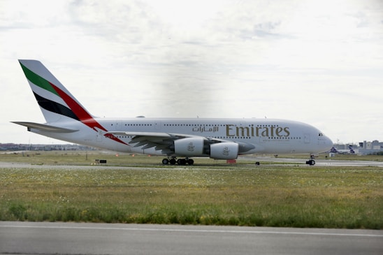 A large Emirates passenger airplane is taxiing on a runway at an airport. The aircraft is painted white with distinctive markings on the tail, featuring red, green, and black stripes. The surrounding area includes grass fields and some distant airport buildings under a cloudy sky.
