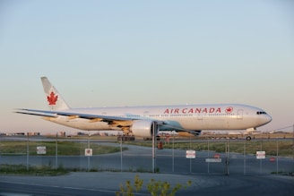 A large commercial airplane with 'Air Canada' branding is parked on an airport runway. The aircraft features a white exterior with a prominent red maple leaf emblem on its tail. The scene is set during daylight with a clear blue sky and a fenced perimeter visible in the foreground. The landscape around the runway shows open grass areas.