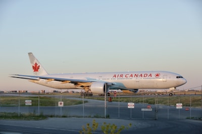 A large commercial airplane with 'Air Canada' branding is parked on an airport runway. The aircraft features a white exterior with a prominent red maple leaf emblem on its tail. The scene is set during daylight with a clear blue sky and a fenced perimeter visible in the foreground. The landscape around the runway shows open grass areas.
