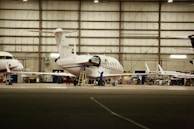 Inside a spacious aircraft hangar, a white jet is being serviced. A person in blue is standing on a ladder near the aircraft, engaged in maintenance work. The hangar is well-lit with several other aircraft visible in the background.