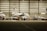 Inside a spacious aircraft hangar, a white jet is being serviced. A person in blue is standing on a ladder near the aircraft, engaged in maintenance work. The hangar is well-lit with several other aircraft visible in the background.