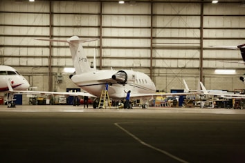 Inside a spacious aircraft hangar, a white jet is being serviced. A person in blue is standing on a ladder near the aircraft, engaged in maintenance work. The hangar is well-lit with several other aircraft visible in the background.
