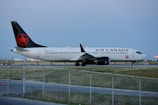 An Air Canada airplane is parked on the runway, surrounded by a fenced off area. The aircraft is primarily white with a black tail featuring a red maple leaf symbol. The runway is bordered by grass and there are some orange cones in the background.