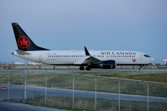 An Air Canada airplane is parked on the runway, surrounded by a fenced off area. The aircraft is primarily white with a black tail featuring a red maple leaf symbol. The runway is bordered by grass and there are some orange cones in the background.