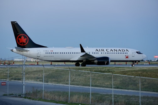 An Air Canada airplane is parked on the runway, surrounded by a fenced off area. The aircraft is primarily white with a black tail featuring a red maple leaf symbol. The runway is bordered by grass and there are some orange cones in the background.
