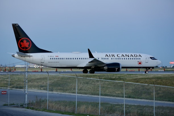 An Air Canada airplane is parked on the runway, surrounded by a fenced off area. The aircraft is primarily white with a black tail featuring a red maple leaf symbol. The runway is bordered by grass and there are some orange cones in the background.
