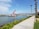 A red and white crane ship is positioned in a body of water near a shoreline. The area is bordered by a paved walkway lined with tall palm trees and green shrubs. The background features distant mountains under a partly cloudy sky.
