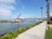 A red and white crane ship is positioned in a body of water near a shoreline. The area is bordered by a paved walkway lined with tall palm trees and green shrubs. The background features distant mountains under a partly cloudy sky.