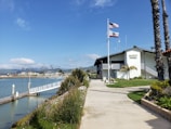 A harbor patrol building is situated beside a body of water with a dock extending out towards the water. The building is white with visible signage and is surrounded by well-maintained greenery and landscaping. Two flags, one American and one Californian, are flying on flagpoles in front of the building. Palm trees and mountains are visible in the background under a clear blue sky.