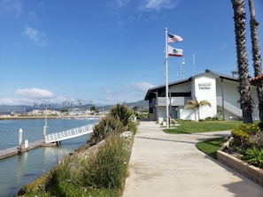 A harbor patrol building is situated beside a body of water with a dock extending out towards the water. The building is white with visible signage and is surrounded by well-maintained greenery and landscaping. Two flags, one American and one Californian, are flying on flagpoles in front of the building. Palm trees and mountains are visible in the background under a clear blue sky.