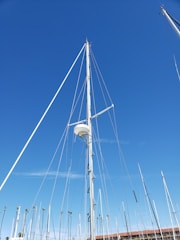 A marina filled with tall sailboat masts against a clear blue sky. The central focus is a prominent white mast with a radar dome attached. Various other masts and rigging wires are visible in the background, with a few palm trees and a low-rise building.